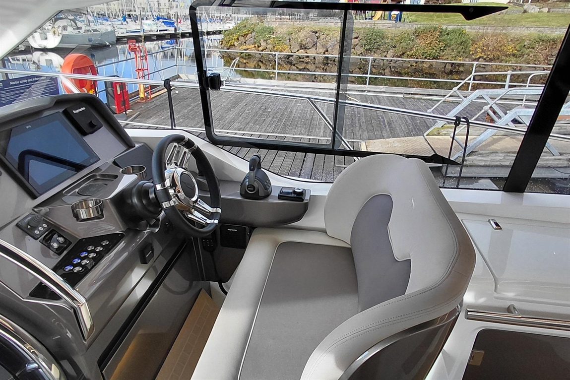 Boat cockpit view featuring a steering wheel, control panel, and comfortable seating, with a scenic marina backdrop.